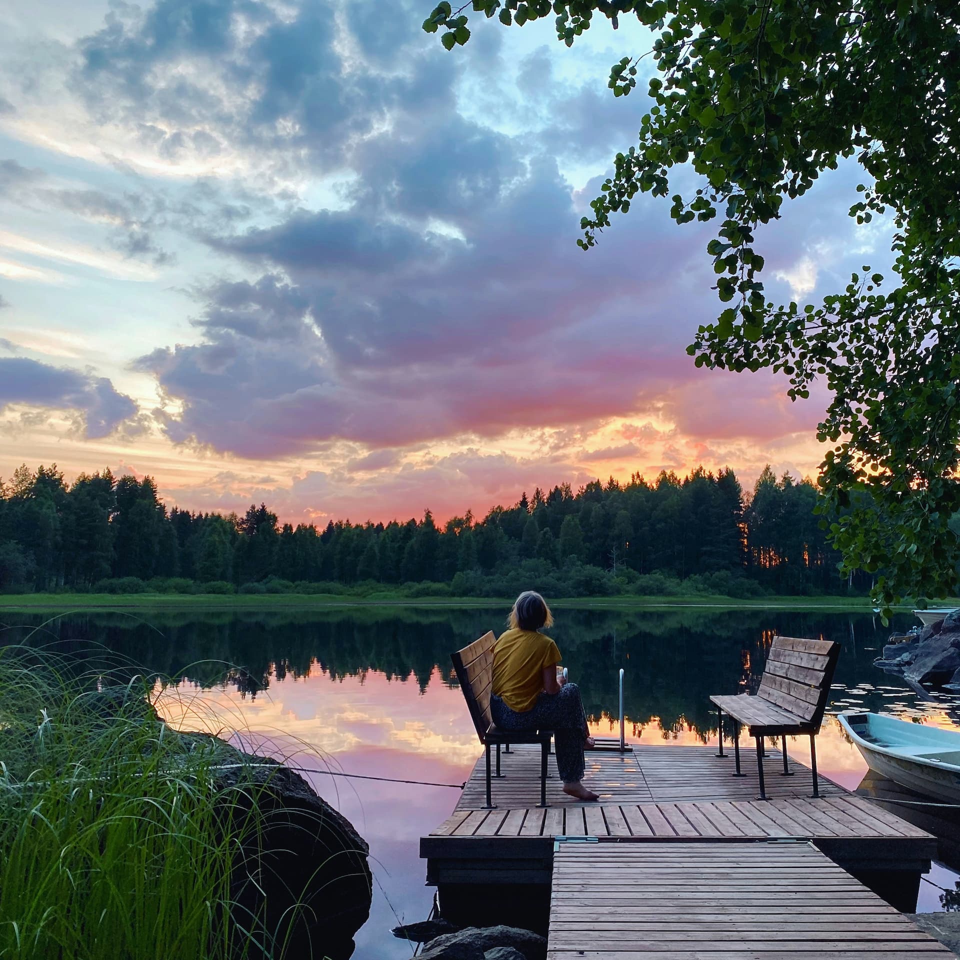 Nainen nauttimassa suomalaisesta järvimaisemasta mökillä, ilta alkaa hämärtyä ja taustalla näkyy auringonlasku kauniissa purppuran värisessä horisontissa